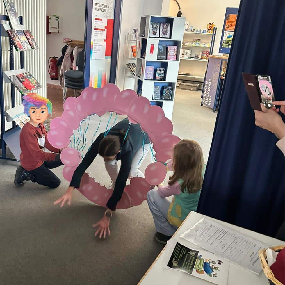 Kinderbuchautorin Alexandra Wagner hechtet während einer Lesung aus "Der Sumpfmumpf und die Hoffnung" durch einen Luftballonring, der von zwei Kindern gehalten wird