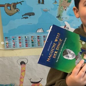 Grundschüler hält das Kinderbuch "Magische Welt der Kinder, Das Geheimnis von Henrietta Handy" in der Hand im Klassenzimmer vor Landkarte