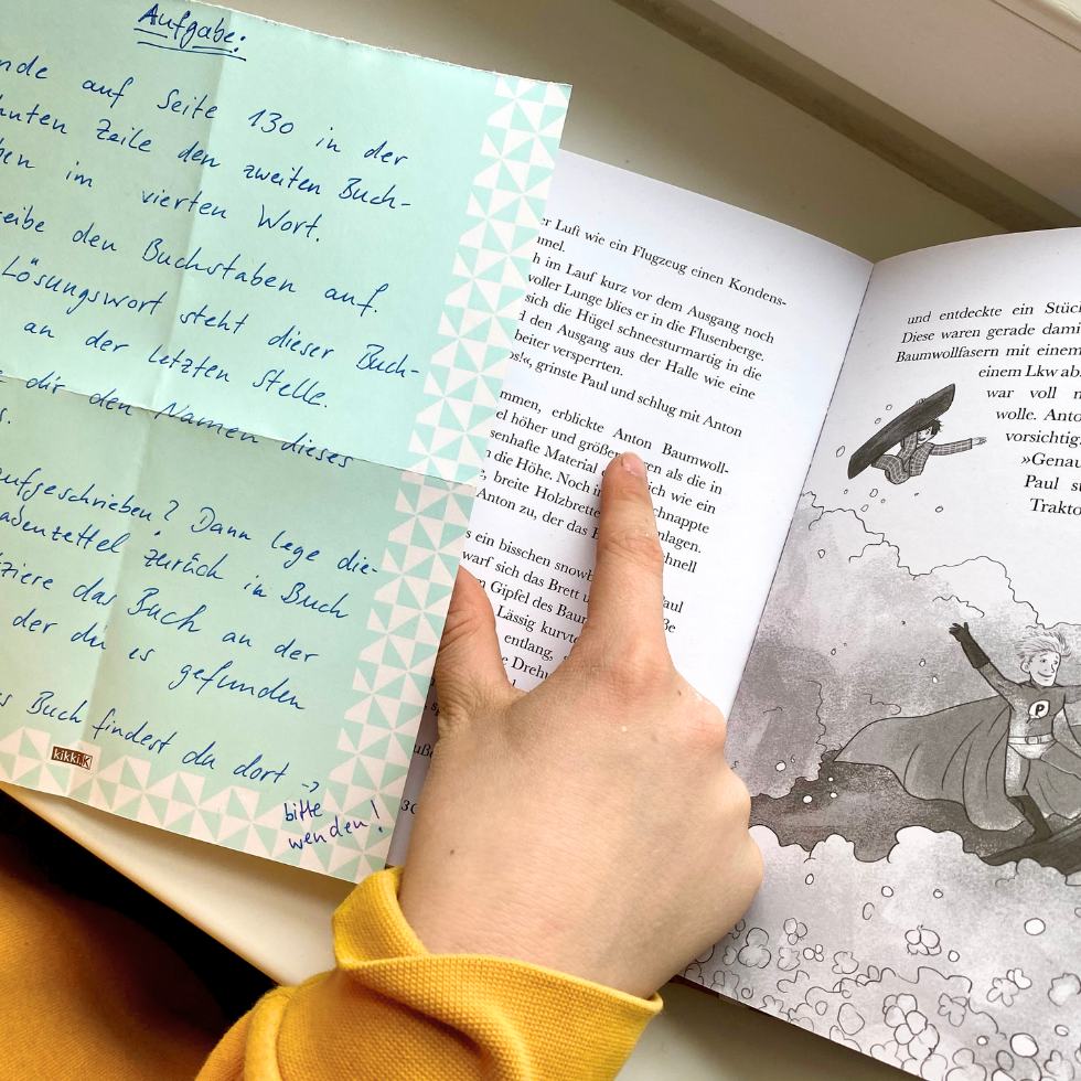 Hand eines Jungen zeigt auf eine Stelle im Buch, daneben der Zettel mit dem Rätsel der Buchstabenjagd