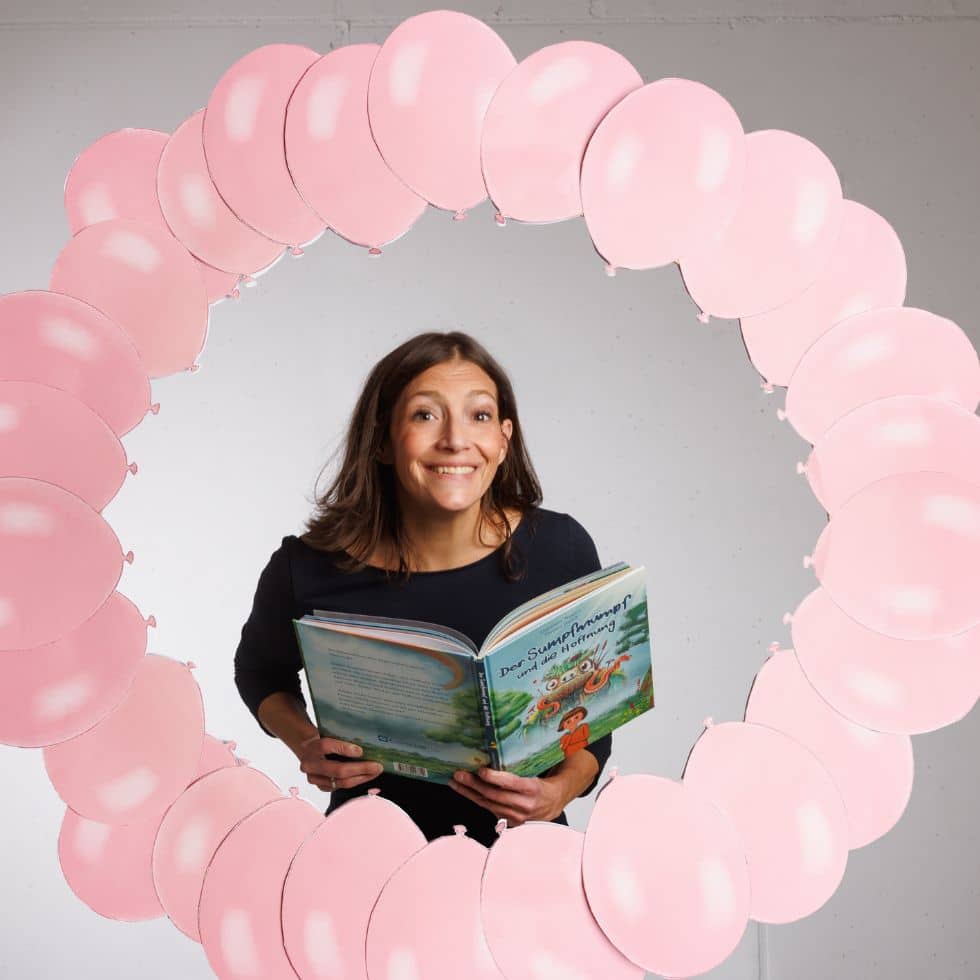 Kinderbuchautorin Alexandra Wagner mit Buch »Der Sumpfnumpf und die Hoffnung« in Händen schaut durch einen Ringa us Luftballons