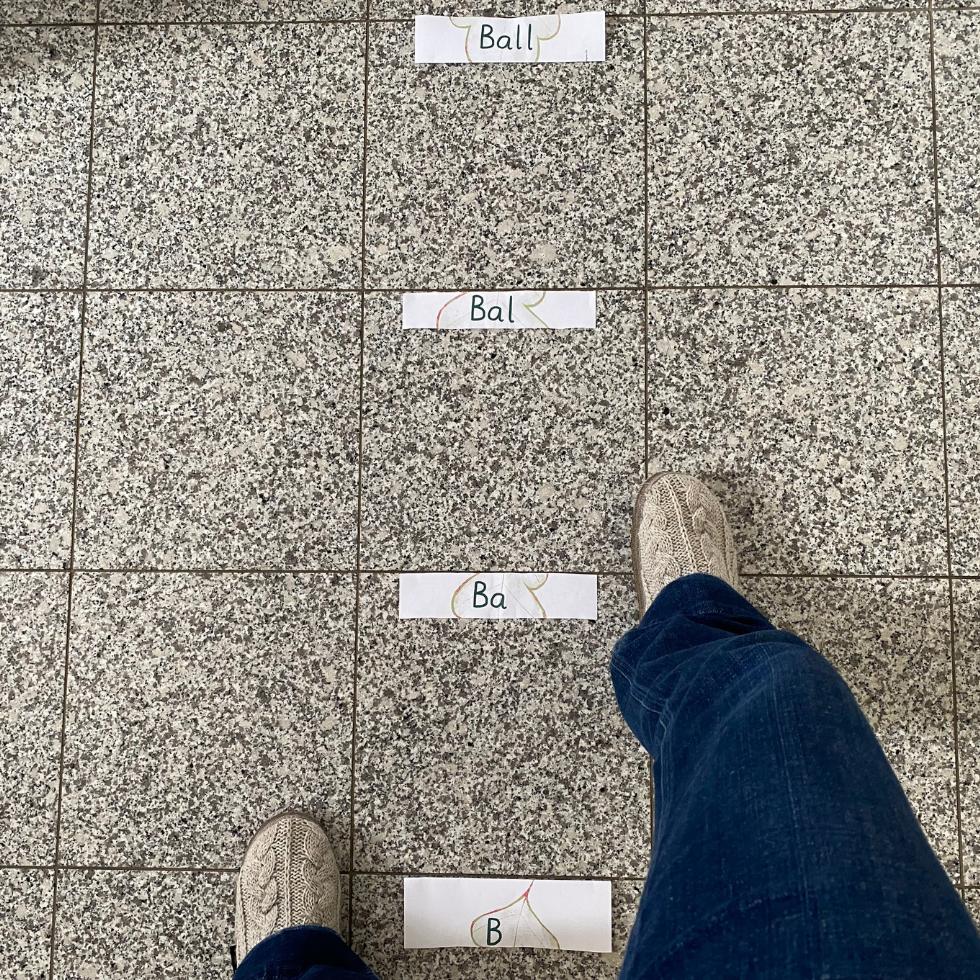 Ausgeschnitten Buchstaben dun Lautverbindungen auf dem Boden und Schrittlänge. Jemand setzt Fuß neben Buchstaben.