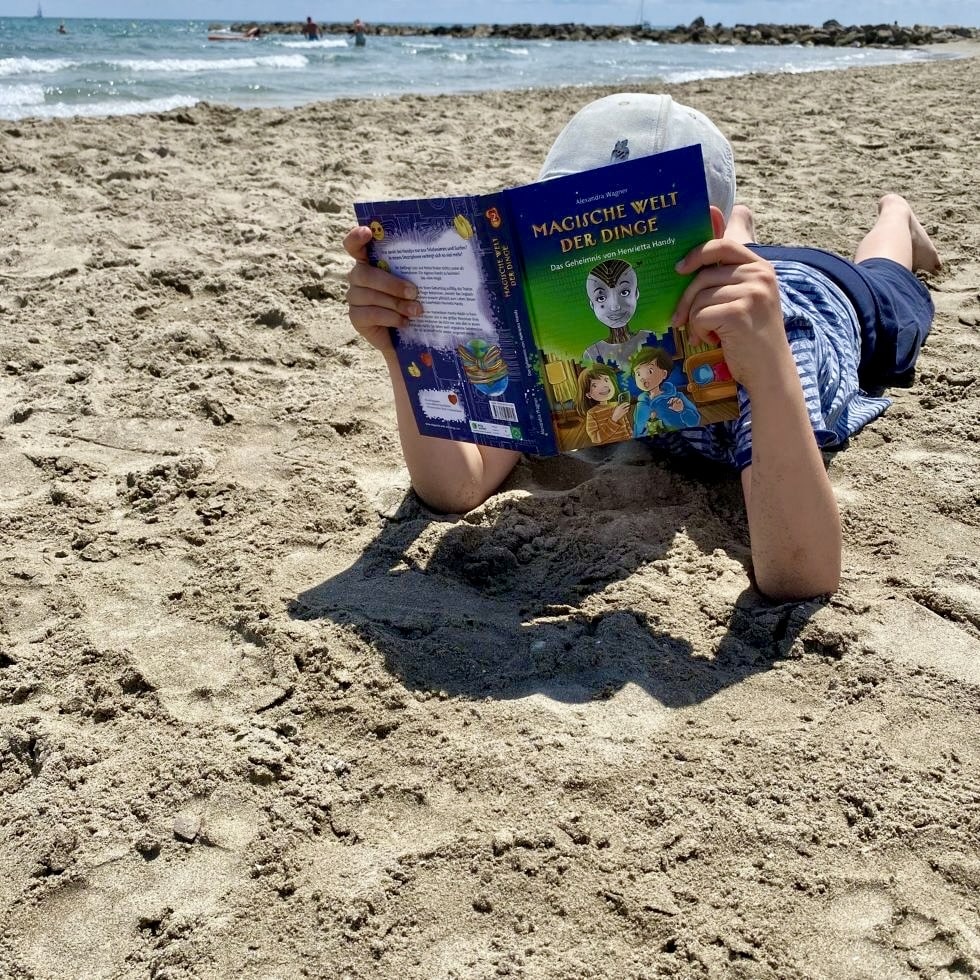 Junge liegt am Sandstrand und vergräbt sein Gesicht im Buch "Magische Welt der Dinge, Das Geheimnis von Henrietta Handy"